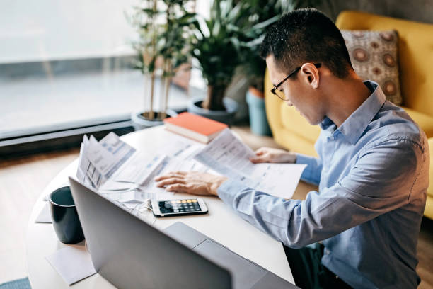 Japanese man with eyeglasses siting on floor in the living room and using smart phone and laptop for managing home finances